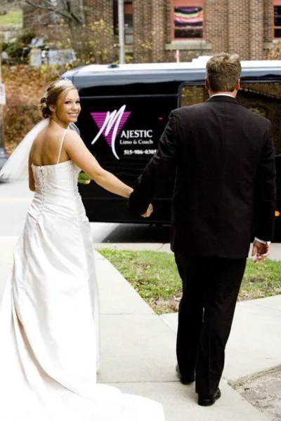 A bride and groom walking hand-in-hand toward a black Majestic Limo & Coach Sprinter van.