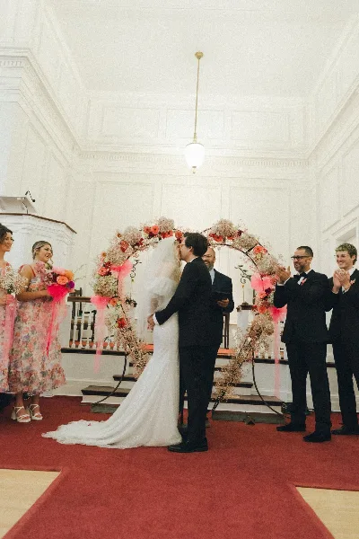 A bride and groom share a kiss during their wedding ceremony under a floral arch, surrounded by guests and an officiant.