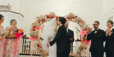 A bride and groom share a kiss during their wedding ceremony under a floral arch, surrounded by guests and an officiant.