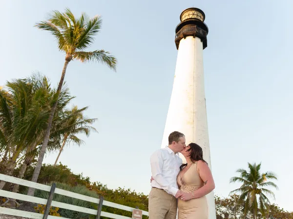 A bride and groom kissing in front of a white lighthouse with palm trees in the background.