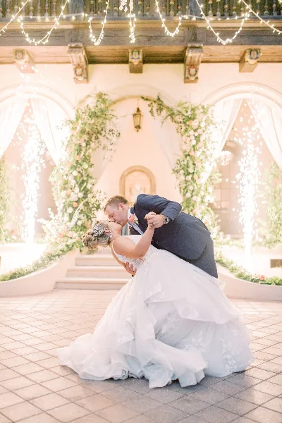 A bride and groom sharing a dip kiss in front of a floral wedding arch and fountain at a venue.