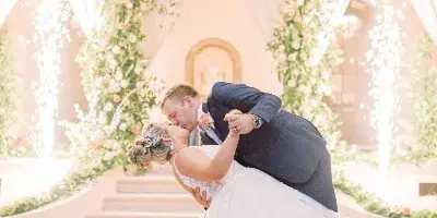 A bride and groom sharing a dip kiss in front of a floral wedding arch and fountain at a venue.