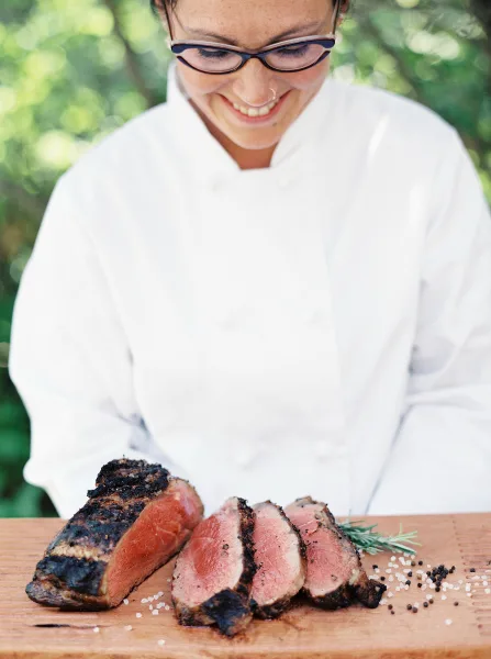 A smiling chef in a white uniform slicing a cooked piece of meat on a wooden cutting board.