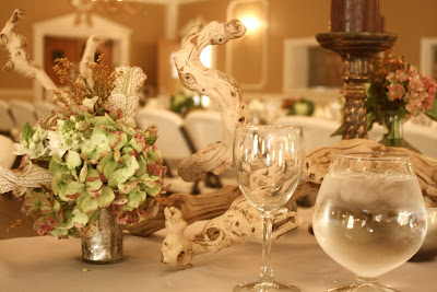 A close-up of a wedding table setting featuring a floral arrangement in a silver vase, driftwood decor, and glassware.