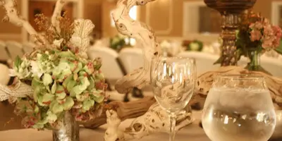 A close-up of a wedding table setting featuring a floral arrangement in a silver vase, driftwood decor, and glassware.