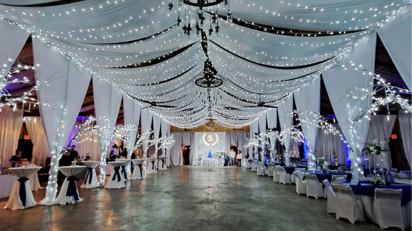 A large event tent interior decorated with white draping, string lights, and set up with tables and chairs for a wedding or party.