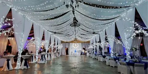 A large event tent interior decorated with white draping, string lights, and set up with tables and chairs for a wedding or party.