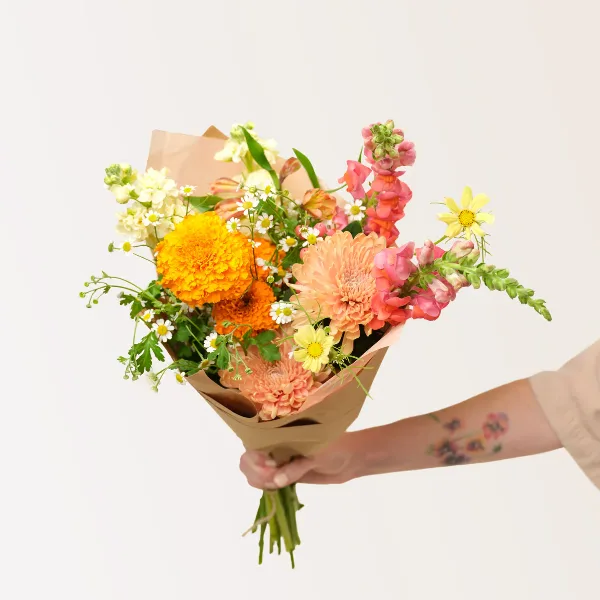 A hand holding a colorful hand-tied bouquet of flowers wrapped in brown paper.
