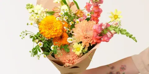 A hand holding a colorful hand-tied bouquet of flowers wrapped in brown paper.