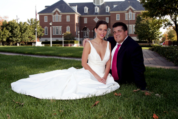 A bride in a white wedding gown and a groom in a dark suit with a pink tie sitting on the grass in front of a large brick building.
