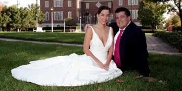 A bride in a white wedding gown and a groom in a dark suit with a pink tie sitting on the grass in front of a large brick building.