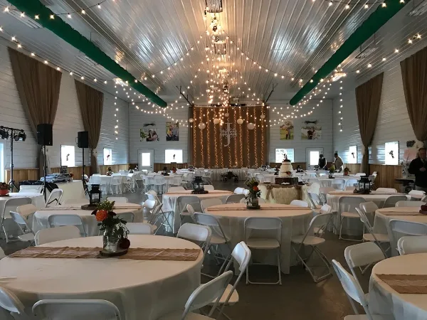 Interior of a rustic barn venue set up for a wedding with round tables, white chairs, and string lights.