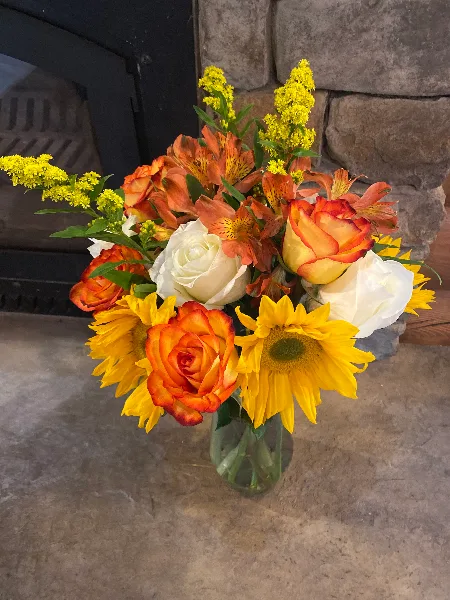 A floral arrangement featuring orange and white roses, alstroemeria, sunflowers, and yellow solidago in a glass vase.