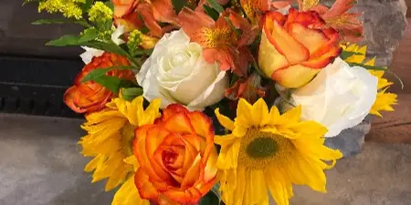 A floral arrangement featuring orange and white roses, alstroemeria, sunflowers, and yellow solidago in a glass vase.