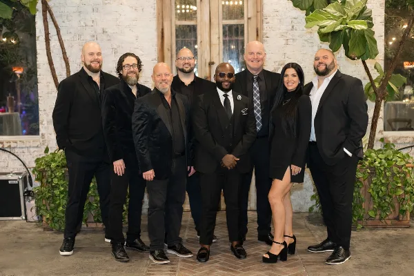 Group of eight musicians in formal black suits and dresses posing for a photo in a venue with white brick walls and greenery.