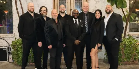Group of eight musicians in formal black suits and dresses posing for a photo in a venue with white brick walls and greenery.