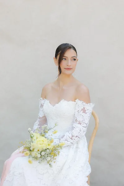 A bride in a white lace gown with a sleek updo and soft makeup, holding a bouquet of yellow roses.