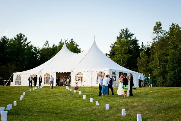 A large white wedding tent set up on a grassy lawn with a bride, groom, and guests standing outside.