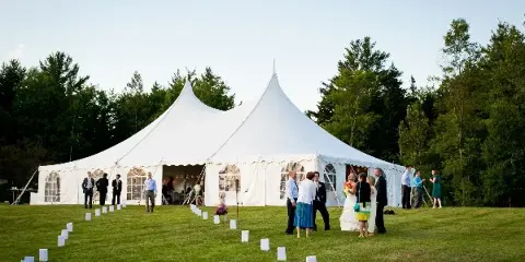 A large white wedding tent set up on a grassy lawn with a bride, groom, and guests standing outside.