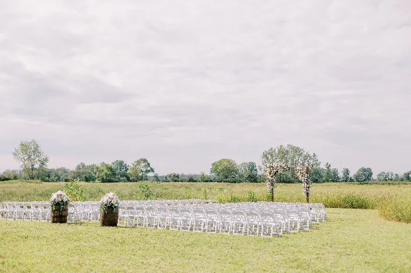 Outdoor wedding ceremony setup with white chairs and a floral arch in a grassy field.