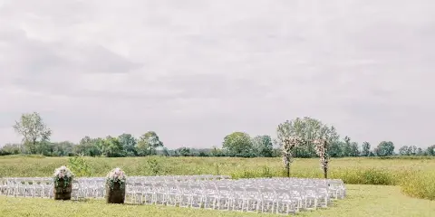 Outdoor wedding ceremony setup with white chairs and a floral arch in a grassy field.