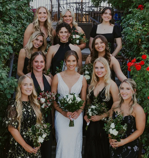A bride in a white wedding dress stands in the center of a group of smiling bridesmaids, all with professionally styled hair and makeup, holding bouquets of white flowers and greenery.