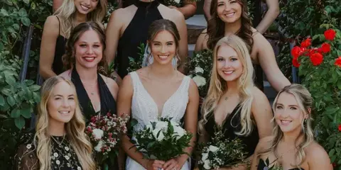 A bride in a white wedding dress stands in the center of a group of smiling bridesmaids, all with professionally styled hair and makeup, holding bouquets of white flowers and greenery.