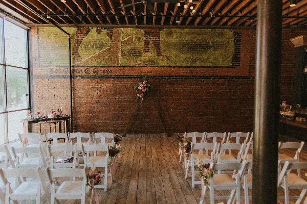Interior of a renovated event space with exposed brick walls and wood plank floors, set up for a wedding ceremony with white chairs and floral decorations.