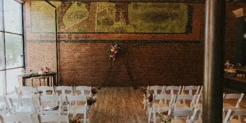 Interior of a renovated event space with exposed brick walls and wood plank floors, set up for a wedding ceremony with white chairs and floral decorations.