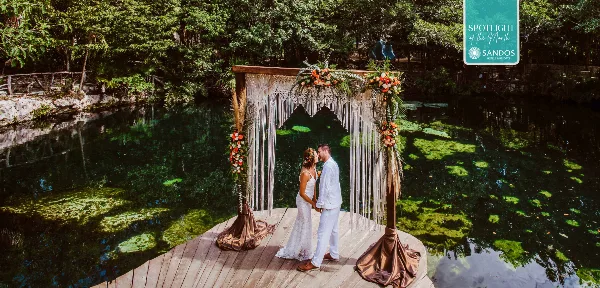 A bride and groom standing under a decorated wedding arch on a wooden platform over a tropical lagoon.