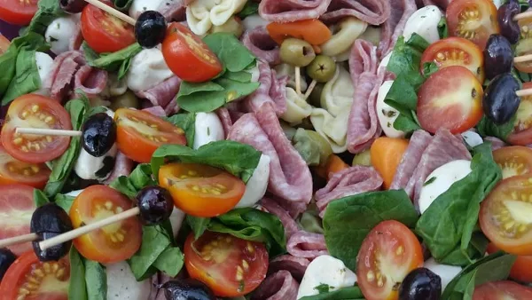 A catering platter featuring tomato and mozzarella skewers, rolled deli meats, olives, and fresh spinach.