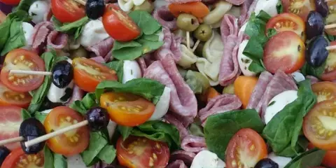 A catering platter featuring tomato and mozzarella skewers, rolled deli meats, olives, and fresh spinach.