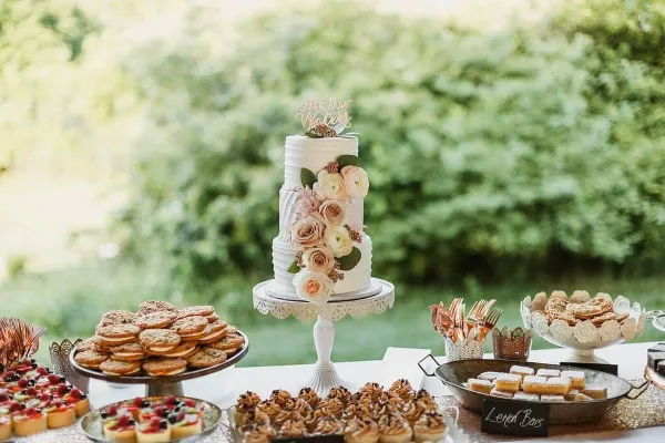 A white wedding cake with floral decorations and a topper on a stand, surrounded by a dessert table with cookies, tarts, and lemon bars.