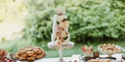 A white wedding cake with floral decorations and a topper on a stand, surrounded by a dessert table with cookies, tarts, and lemon bars.