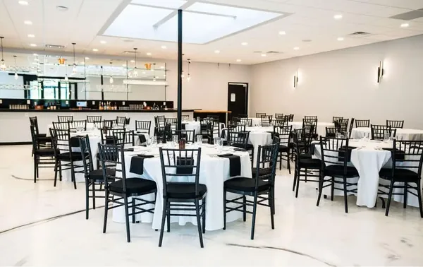 Event venue dining room set with round tables, white tablecloths, black chairs, and a bar in the background.
