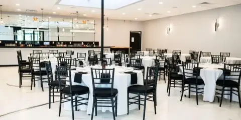 Event venue dining room set with round tables, white tablecloths, black chairs, and a bar in the background.