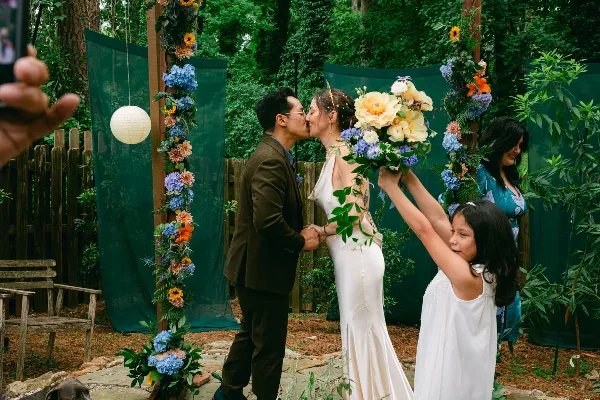 A bride and groom kissing during an outdoor wedding ceremony with a floral arch and guests watching.
