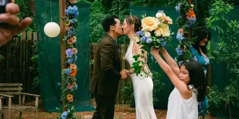 A bride and groom kissing during an outdoor wedding ceremony with a floral arch and guests watching.