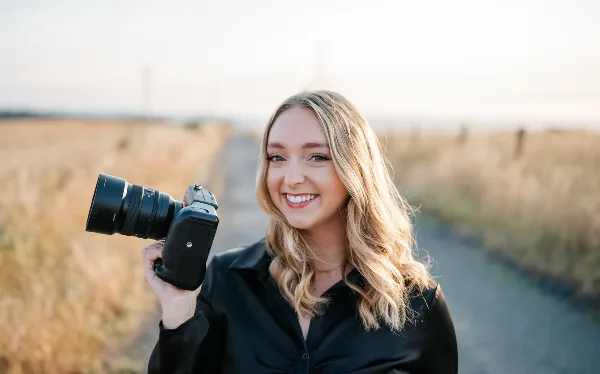 A smiling woman with blonde hair holding a professional camera in a field.