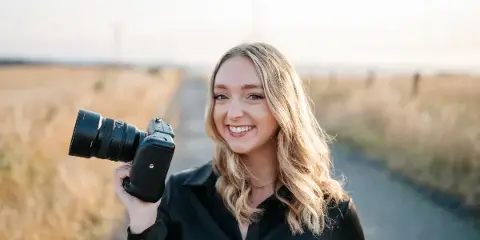 A smiling woman with blonde hair holding a professional camera in a field.
