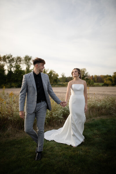 A bride and groom holding hands and walking in a field during a wedding photoshoot.