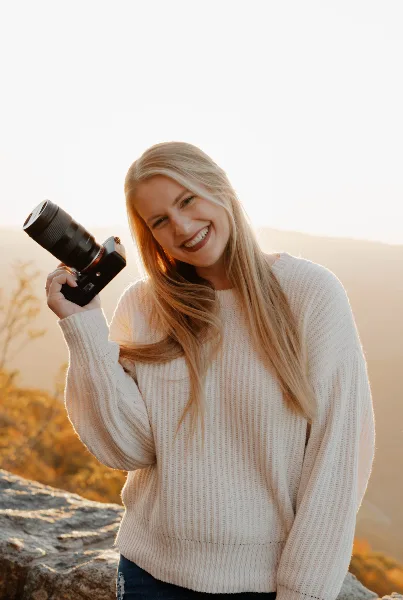 A smiling blonde woman holding a professional camera with a lens against a mountain sunset background.