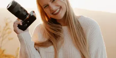 A smiling blonde woman holding a professional camera with a lens against a mountain sunset background.