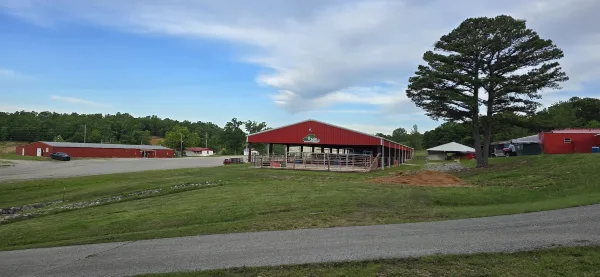 Exterior view of a fairground facility with red buildings, an open-sided arena, and grassy grounds under a partly cloudy sky.