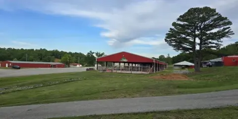 Exterior view of a fairground facility with red buildings, an open-sided arena, and grassy grounds under a partly cloudy sky.