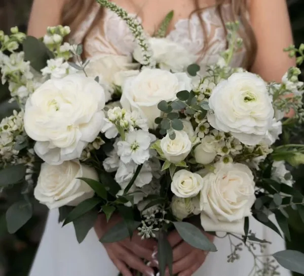 A bride in a white lace dress holding a large bouquet of white roses, ranunculus, and eucalyptus.