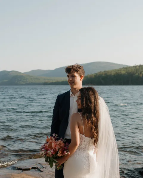 A bride and groom standing together on a rocky shoreline with a lake and forested mountains in the background.