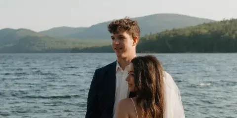 A bride and groom standing together on a rocky shoreline with a lake and forested mountains in the background.