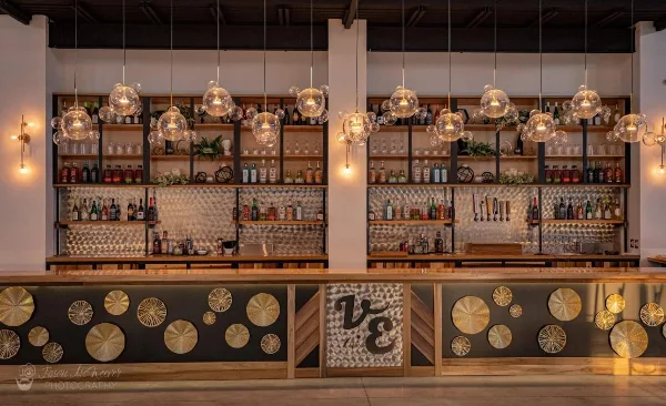 Interior view of the Vibrant Event Center bar featuring glass pendant lights, stocked shelves, and a decorative front panel with the 'VE' logo.