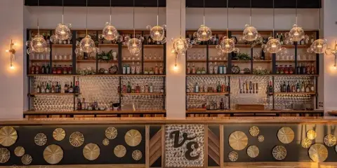 Interior view of the Vibrant Event Center bar featuring glass pendant lights, stocked shelves, and a decorative front panel with the 'VE' logo.